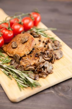 Tasty pork meat with grilled mushrooms next to oregano branch on wooden board. Cherry tomatoes are in blurred backgorund