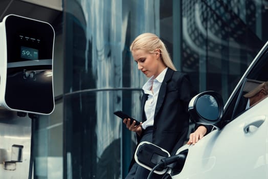 Businesswoman wearing black suit using smartphone, leaning on electric car recharge battery at charging station in city residential building with condos and apartment. Progressive lifestyle concept.