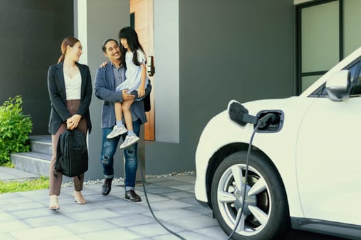 Progressive young parents and daughter with electric vehicle and home charging station. Green and clean energy from electric vehicles for healthy environment. Eco power from renewable source at home.