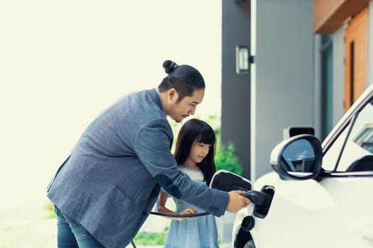 Progressive father and daughter plugs EV charger from home charging station to electric vehicle. Future eco-friendly car with EV cars powered by renewable source of clean energy.