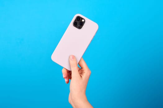 Hand of unrecognizable person showing back side of modern silver smartphone against blue backdrop in studio