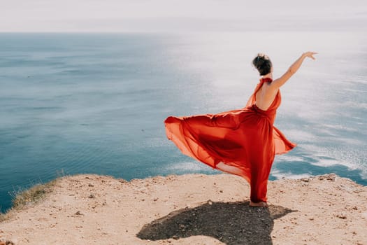 Side view a Young beautiful sensual woman in a red long dress posing on a rock high above the sea during sunrise. Girl on the nature on blue sky background. Fashion photo.