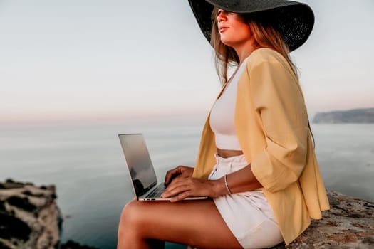 Successful business woman in yellow hat working on laptop by the sea. Pretty lady typing on computer at summer day outdoors. Freelance, travel and holidays concept.