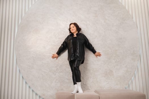 An adult woman in black leather clothes poses against a gray studio wall.