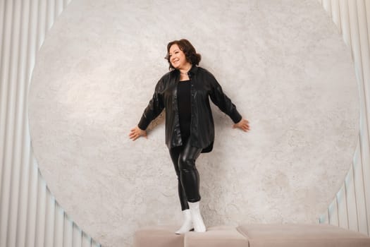 An adult woman in black leather clothes poses against a gray studio wall.