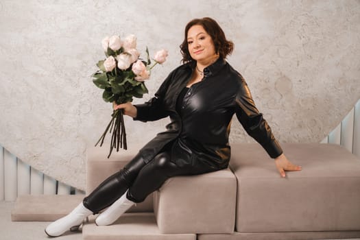 a stylish adult woman in black leather clothes is sitting with a bouquet of pink roses in the interior.