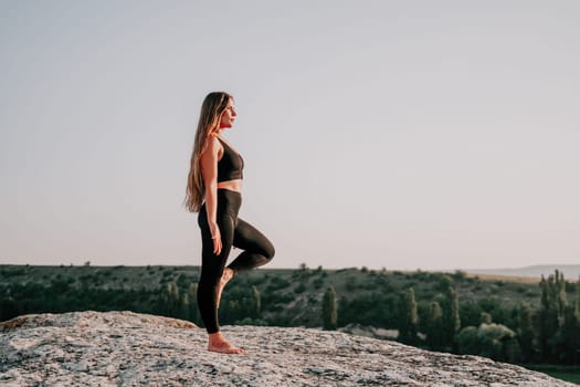 Well looking middle aged woman with long hair, fitness instructor in leggings and tops doing stretching and pilates on the rock near forest. Female fitness yoga routine concept. Healthy lifestyle.