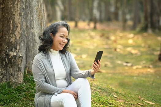 Positive middle aged woman using smart phone under tree on green grass at sunny beautiful day.