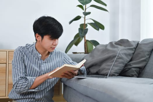 Pleasant asian man sitting on floor and reading book, spending leisure time at home. Leisure and people concept.