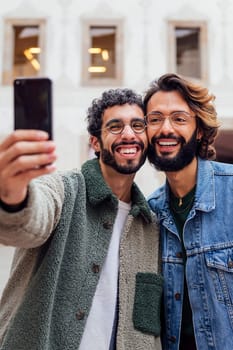 vertical portrait of a happy couple of gay men laughing taking a selfie photo with a mobile phone in the street, concept of urban lifestyle and love between people of the same sex