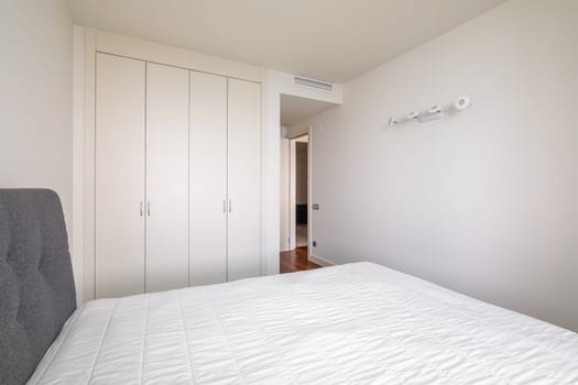Modern bright bedroom with a bed and built-in wardrobe and an open door to the corridor. Minimalistic but cozy design concept. Hotel or rental apartment design.