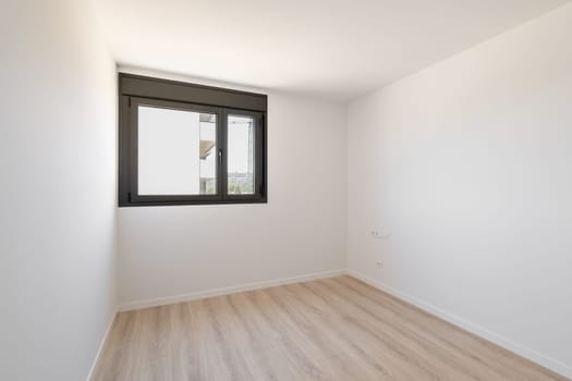 Small empty room without furniture. The floor is new light wood laminate. A window made of black plastic for airing the room. Daylight enters through the frosted glass in the window