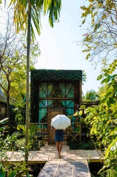 Asian women walk with umbrella at a wooden cottage surrounded by palm trees and a vegetable garden in the countryside. cabin in tropical rainforest