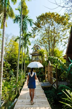 Asian women walk with umbrella at a wooden cottage surrounded by palm trees and a vegetable garden in the countryside. cabin in tropical rainforest