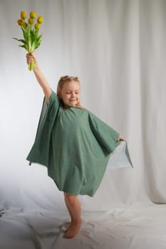 Portrait of a little blonde girl with bouquet of spring yellow flowers on a light background. Child in green dress holding a bouquet of tulips in hands. Spring concept