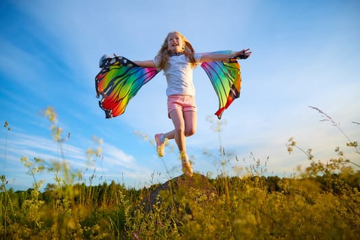 Pretty blonde girl with bright butterfly wings having fun in meadow on natural landscape with grass and flowers on sunny summer day. Portrait of teenage child in spring season outdoors on field