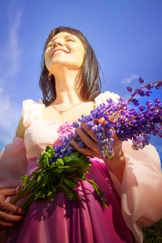 Beautiful girl in lush pink ball gown in green field during blooming of flowers and blue sky on background. Model posing on nature landscape as princess from fary tale