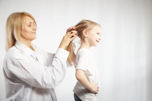 Close portrait of blonde mother and daughter where mom braids hair and makes ponytail and hairstyle for her girl on white background in studio. The concept of love, friendship, caring in family