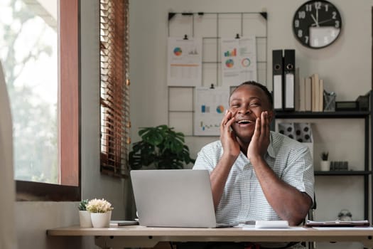 Smiling black man using laptop at home in living room. Happy mature businessman send email and working at home. African american freelancer typing on computer with paperworks and documents on table...