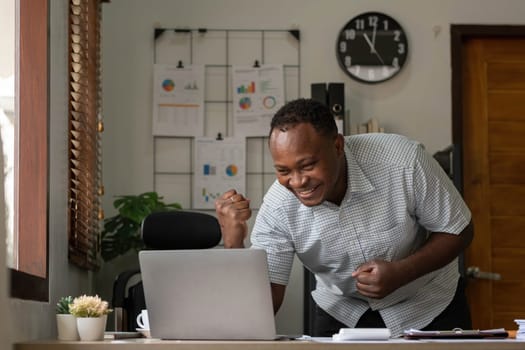 Smiling black man using laptop at home in living room. Happy mature businessman send email and working at home. African american freelancer typing on computer with paperworks and documents on table...