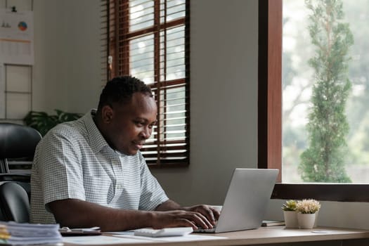 Smiling black man using laptop at home in living room. Happy mature businessman send email and working at home. African american freelancer typing on computer with paperworks and documents on table...