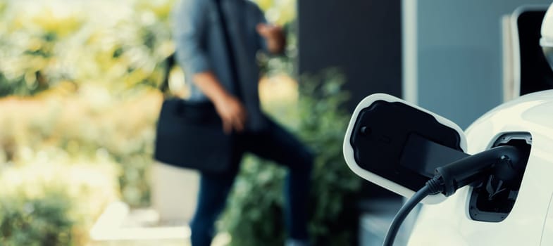 Focus electric car charging at home charging station with blurred progressive man walking in the background. Electric car using renewable clean for eco-friendly concept.