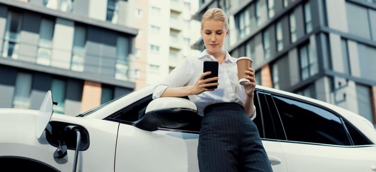 Focus businessman using phone, leaning on electric vehicle, holding coffee with blurred city residential condo buildings in background as progressive lifestyle by renewable and sustainable EV car.