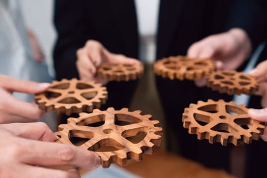 Closeup hand holding wooden gear by businesspeople wearing suit for harmony synergy in office workplace concept. Group of people hand making chain of gears into collective form for unity symbol.