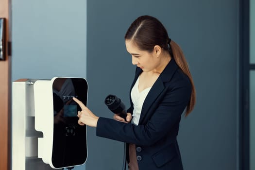 Asian woman holding and pointing an EV plug, a home charging station providing a sustainable power source for electric vehicles. Alternative energy for progressive lifestyle.