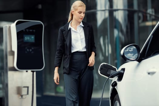 Progressive businesswoman wearing suit with electric car recharging at public parking car charging station at modern city center. Eco friendly rechargeable car powered by alternative clean energy.