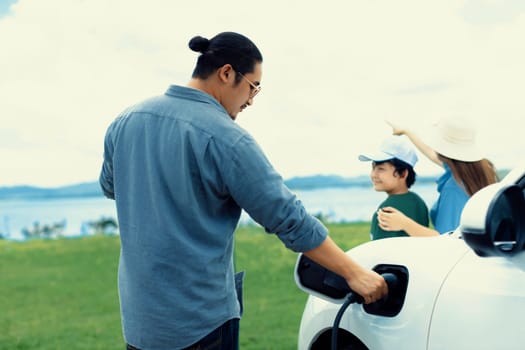 Concept of progressive happy family enjoying their time at green field and lake with electric vehicle. Electric vehicle driven by clean renewable from eco-friendly power sauce.