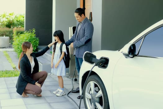 Progressive young parents and daughter with electric vehicle and home charging station. Green and clean energy from electric vehicles for healthy environment. Eco power from renewable source at home.