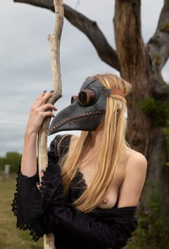 A captivating image of a nude woman donning a black crow mask, standing by an old dry tree in the midst of nature's tranquility.