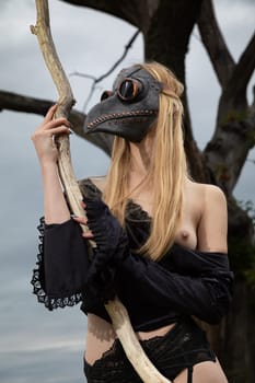 A captivating image of a nude woman donning a black crow mask, standing by an old dry tree in the midst of nature's tranquility.