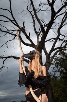 A captivating image of a nude woman donning a black crow mask, standing by an old dry tree in the midst of nature's tranquility.