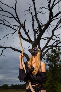 A captivating image of a nude woman donning a black crow mask, standing by an old dry tree in the midst of nature's tranquility.