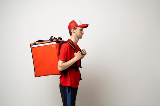 Side view of young courier with curly hair in red uniform with red thermo bag on white background. Food delivery service