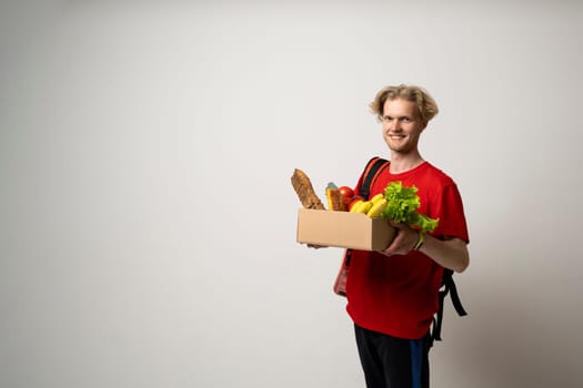 Happy delivery man in red uniform with a box full of groceries over white background. Express delivery, food delivery, online shopping concept