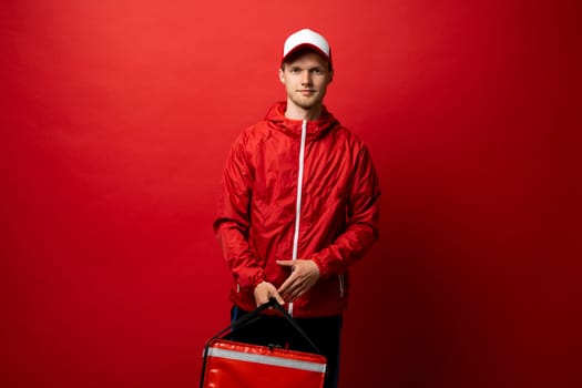 Delivery Service Concept. Portrait of confident smiling male courier wearing red uniform and thermo backpack bag on red studio background