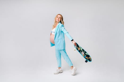 a pregnant girl in turquoise clothes with a skateboard in her hands on a gray background.