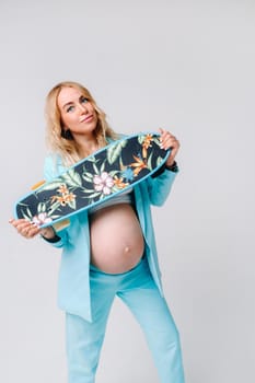 a pregnant girl in turquoise clothes with a skateboard in her hands on a gray background.