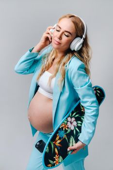 A pregnant girl in a turquoise suit with a skateboard in her hands and headphones stands on a gray background.