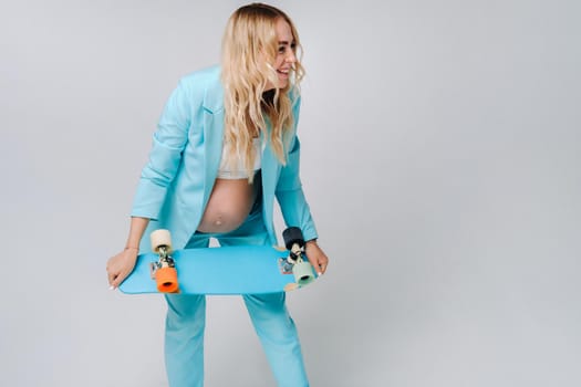 a pregnant girl in turquoise clothes with a skateboard in her hands on a gray background.