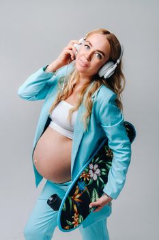 A pregnant girl in a turquoise suit with a skateboard in her hands and headphones stands on a gray background.