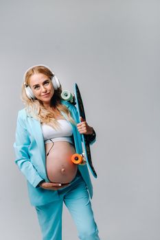 A pregnant girl in a turquoise suit with a skateboard in her hands and headphones stands on a gray background.