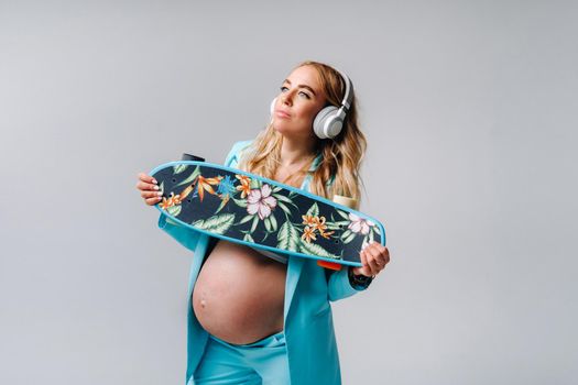 A pregnant girl in a turquoise suit with a skateboard in her hands and headphones stands on a gray background.