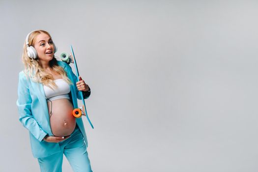 A pregnant girl in a turquoise suit with a skateboard in her hands and headphones stands on a gray background.
