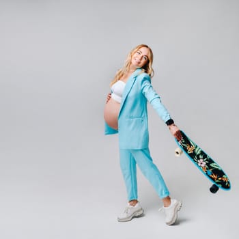 a pregnant girl in turquoise clothes with a skateboard in her hands on a gray background.