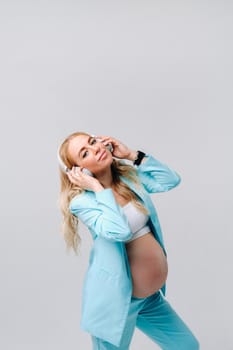 a pregnant woman in a turquoise suit with headphones stands and listens to music on a gray background.