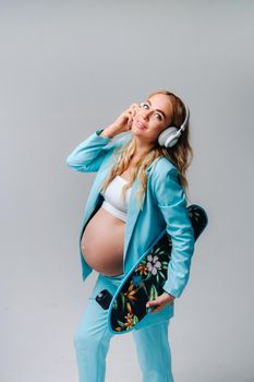 A pregnant girl in a turquoise suit with a skateboard in her hands and headphones stands on a gray background.